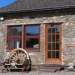 View of the entrance to the but-n-ben stone cottage with a wagin wheel and wooden bench to the side of the front door